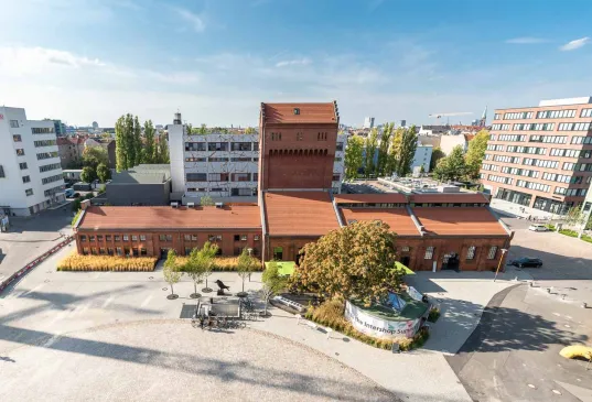 Aerial view of the EUREF Campus in Berlin with historic brick buildings and modern office complexes in the background. In the foreground, there are trees and a paved square
