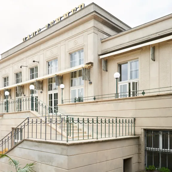 Bright building with outdoor terrace, tables and chairs, surrounded by trees.