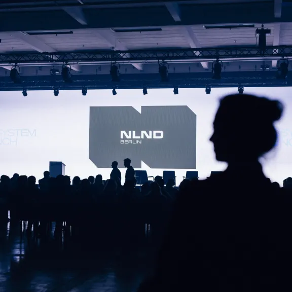 Schattenriss einer Person vor einer beleuchteten Konferenzbühne mit Publikum.