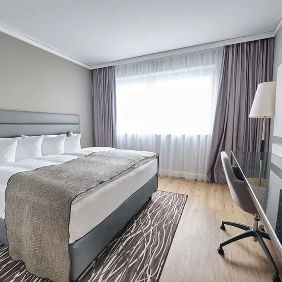 Modernes Hotelzimmer mit Doppelbett, Schreibtisch, Stuhl und großem Fenster mit Vorhängen.