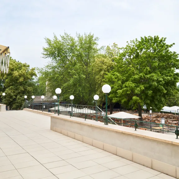 Spacious terrace overlooking trees, lamps and a river in daylight.