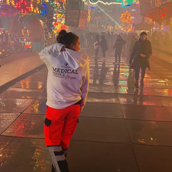 Sanitäterin mit weißer Jacke und roter Hose läuft nachts über einen bunten Jahrmarkt.