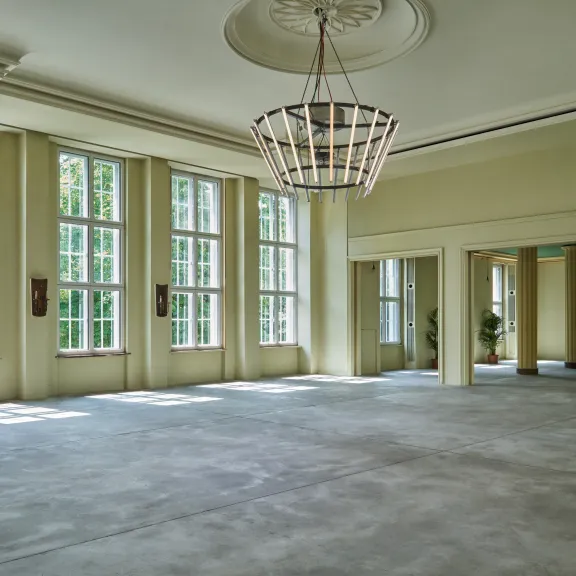 Großer, leerer Saal mit hohen Fenstern und Kronleuchter, viel Tageslicht.