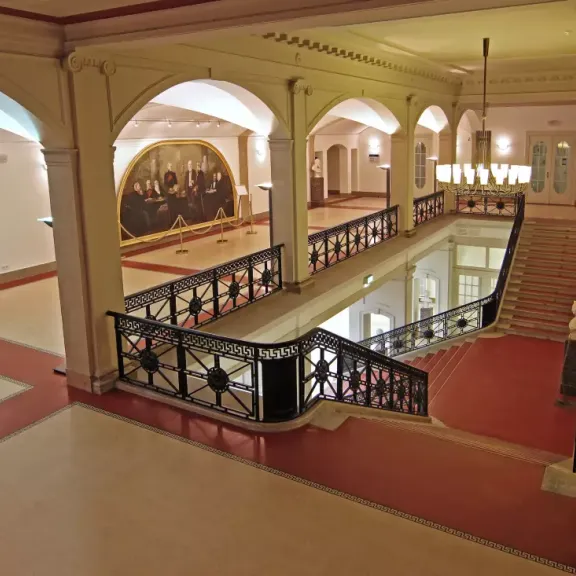 Elegantes Treppenhaus mit rotem Teppich, Gemälde und klassischen Säulen.