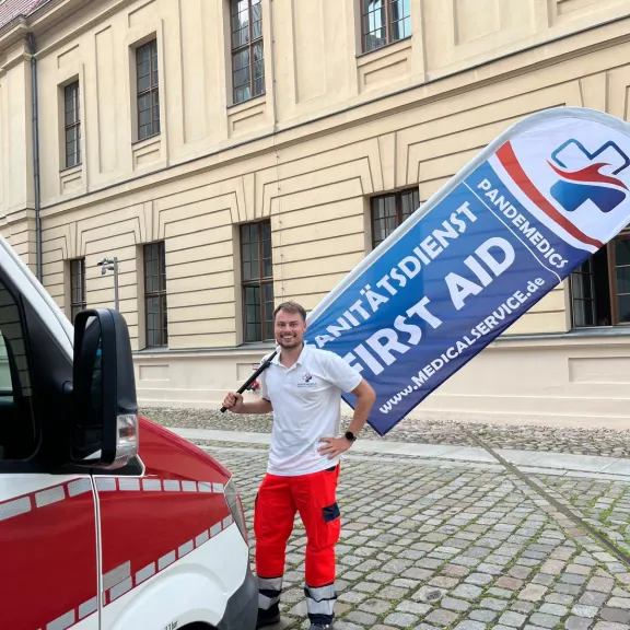 Sanitäter steht neben einem Krankenwagen und hält eine Fahne mit „First Aid“-Aufschrift.