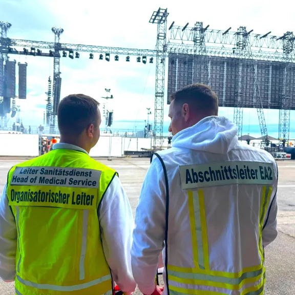 Zwei Sanitäter im weißen Outfit stehen vor einer großen Konzertbühne im Freien.