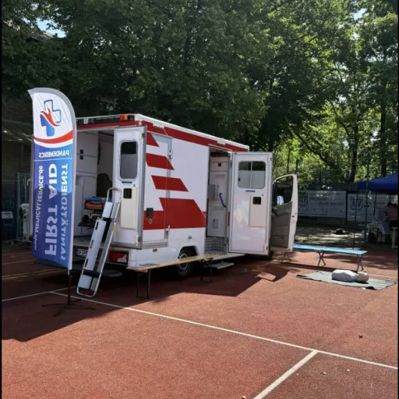 Mobiler Rettungswagen auf rotem Sportplatz, Bäume im Hintergrund.