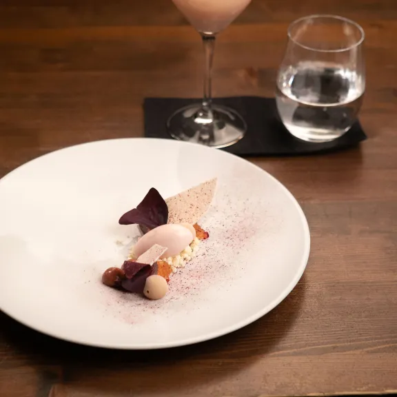 Elegantes Dessert auf weißem Teller, daneben Cocktail und Wasserglas auf dunklem Holztisch.