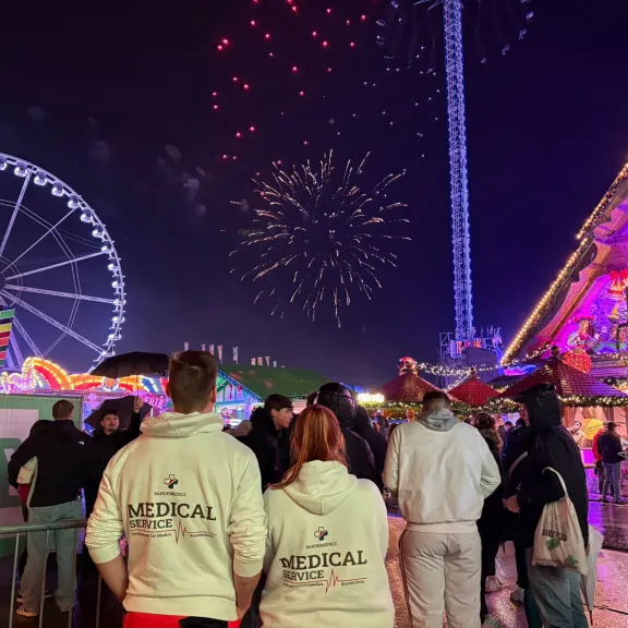 Menschenmenge auf einem beleuchteten Jahrmarkt mit Riesenrad und Feuerwerk am Nachthimmel.