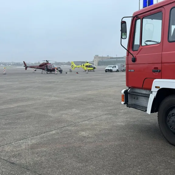 Rotes Feuerwehrfahrzeug, zwei Hubschrauber auf Flugfeld bei grauem Himmel.