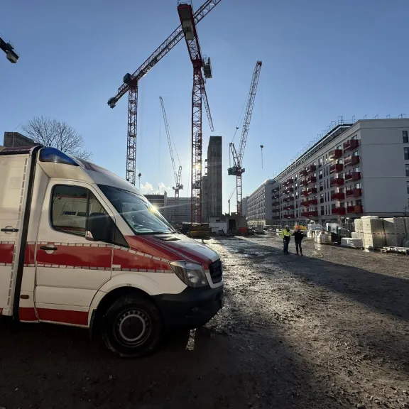 Krankenwagen vor Baustelle mit mehreren Baukränen und Wohngebäuden im Hintergrund.