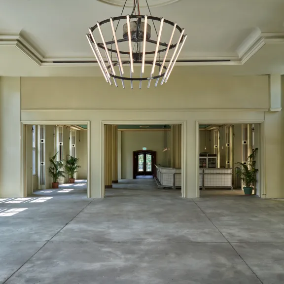 Leerer, heller Raum mit großen Fenstern, Kronleuchter und Pflanzen in den Ecken.