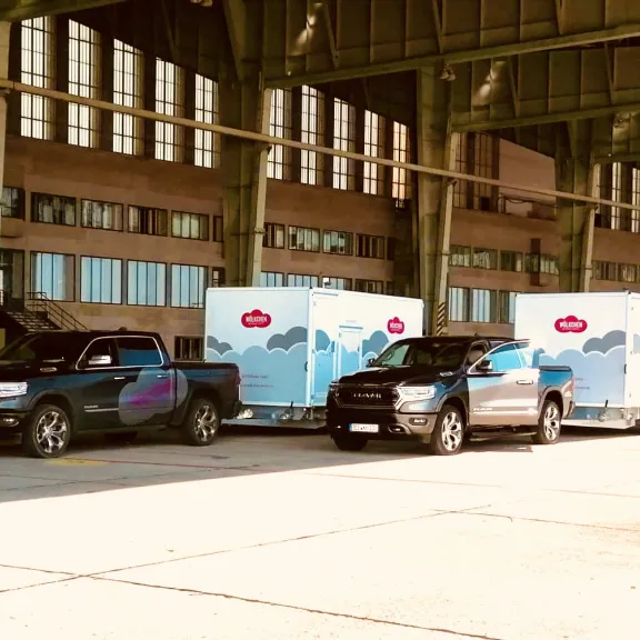 Hier stehen zwei Pick Up, mit jeweils einem WC-Wagen unter einem großen Vordach eines Hangars.