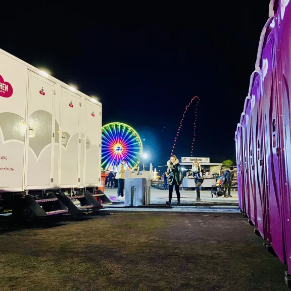 Hier sind auf der rechten Seite mehrere, pinke Wölkchen WC-Kabinen zu sehen. Auf der linken Seite steht ein mobiler WC-Wagen mit dem Logo, einer pinken Wolke und weißem Schriftzug "Wölkchen-Die frische Toilette". im Hintergrund steht ein sich drehendes Riesenrad.