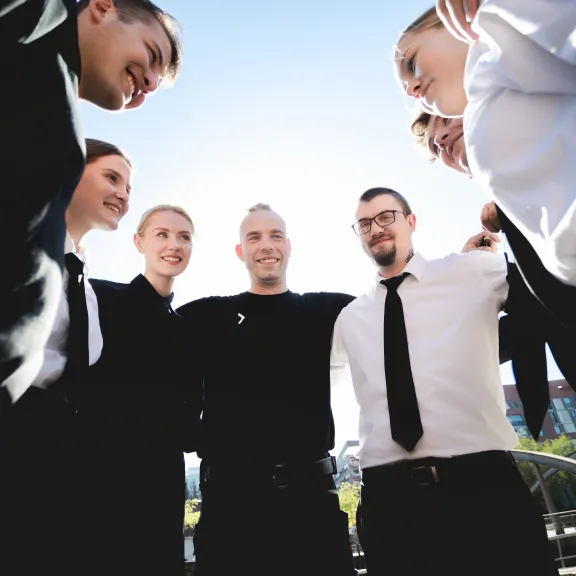 Sechs Menschen stehen lachend im Kreis, sonniger Tag, Teamstimmung.