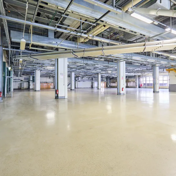 Leere Industriehalle mit Betonboden und sichtbaren Deckenrohren.