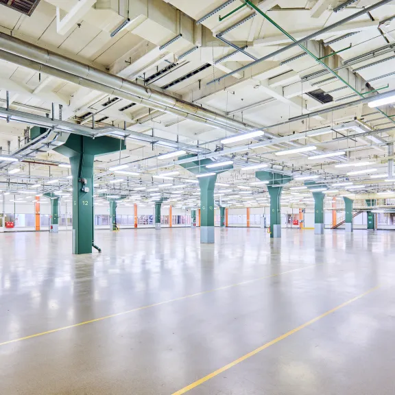 Große, leere Industriehalle mit grünen Säulen und hellen Deckenleuchten.