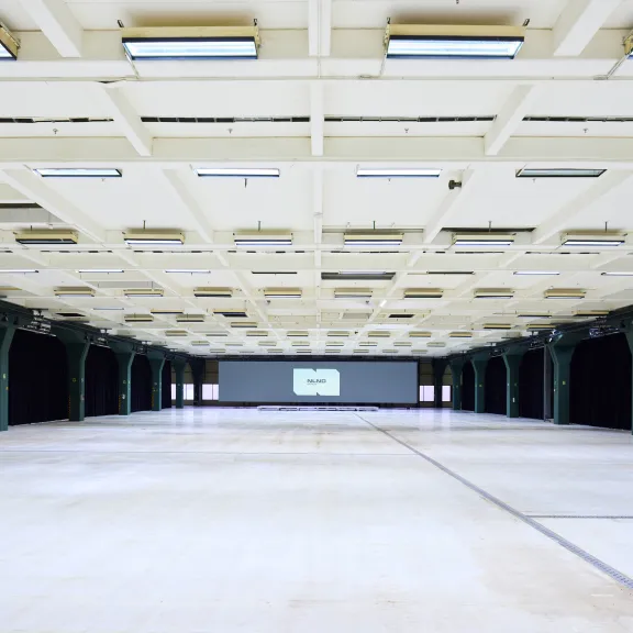 Leere, helle Industriehalle mit großen Deckenlampen und schwarzem Vorhang am Rand.