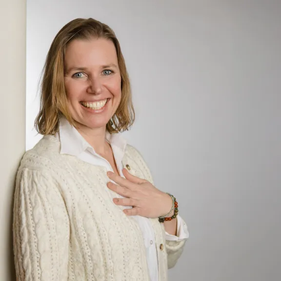 Frau mit hellem Pullover lächelt und lehnt an einer weißen Wand.
