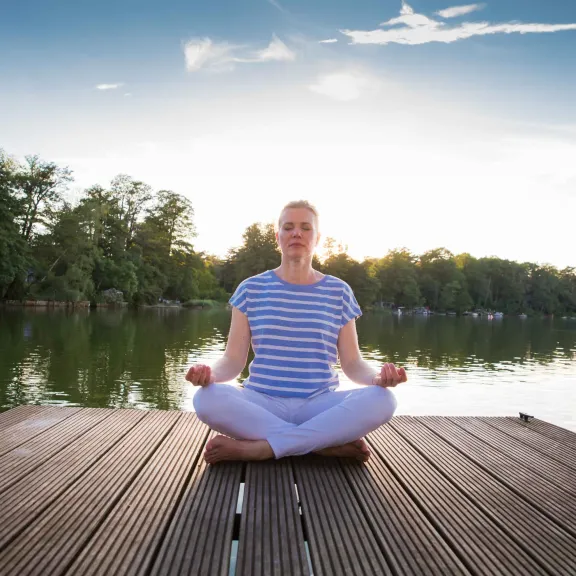Person meditiert im Schneidersitz auf einem Steg am See bei Sonnenuntergang.