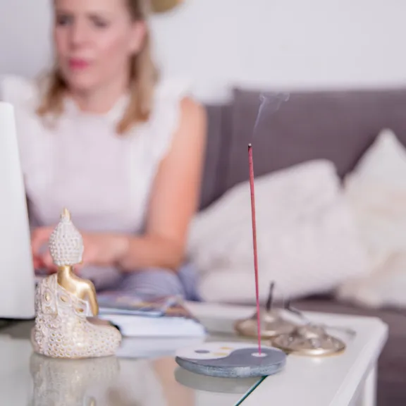Frau sitzt am Laptop auf dem Sofa, auf dem Tisch eine Buddha-Statue und brennendes Räucherstäbchen.
