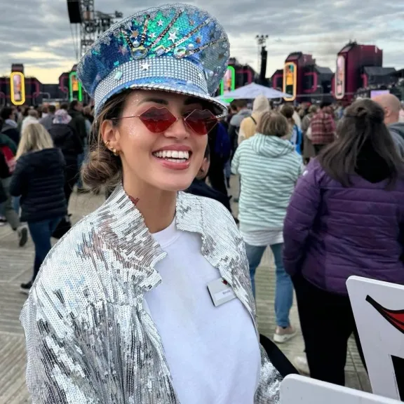 Frau lächelt in die Kamera, trägt glitzernden Hut und silberne Glitzerjacke, im Hintergrund viele Menschen – Eventpersonal und Promotionteam im Einsatz.