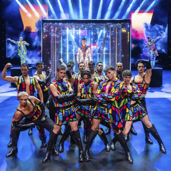 Tanzgruppe in bunten Kostümen auf einer hell erleuchteten Theaterbühne.