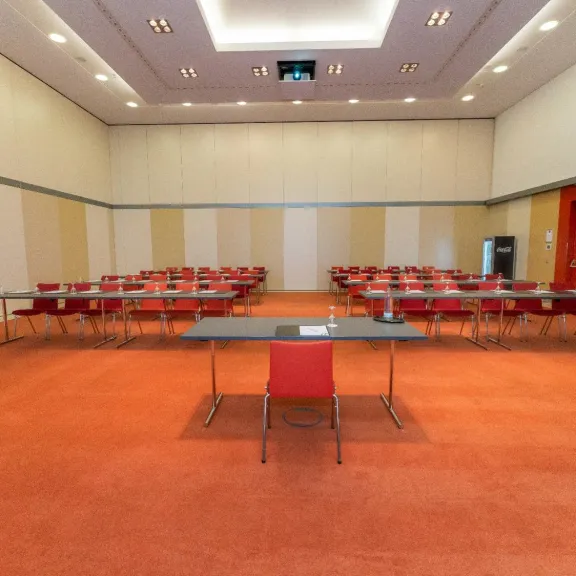 Leerer Konferenzraum mit roten Stühlen und Tischen, rote Tür rechts, Teppichboden.