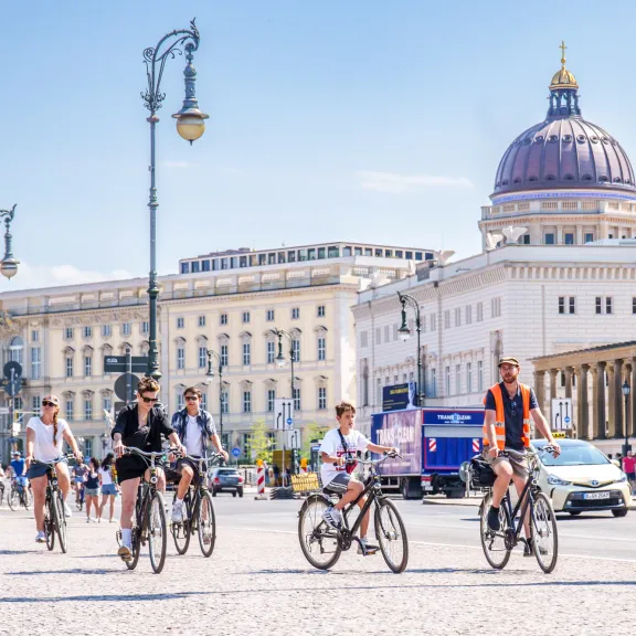 Mehrere Menschen fahren bei sonnigem Wetter Fahrrad vor historischen Gebäuden.