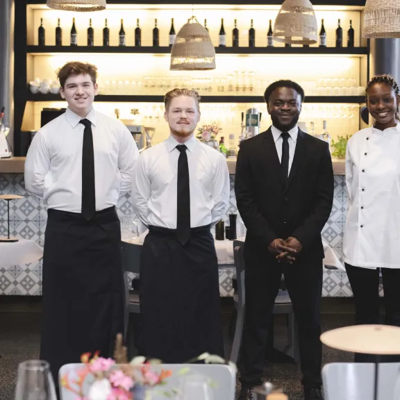 Vier Restaurantmitarbeiter stehen lächelnd vor der Bar im Gastraum.