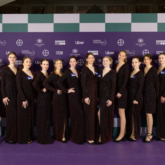 14 Frauen in identischen glitzernden Kleidern stehen auf einem lila Teppich vor einer Sponsorenwand – Eventpersonal und Promotionteam im Einsatz.
