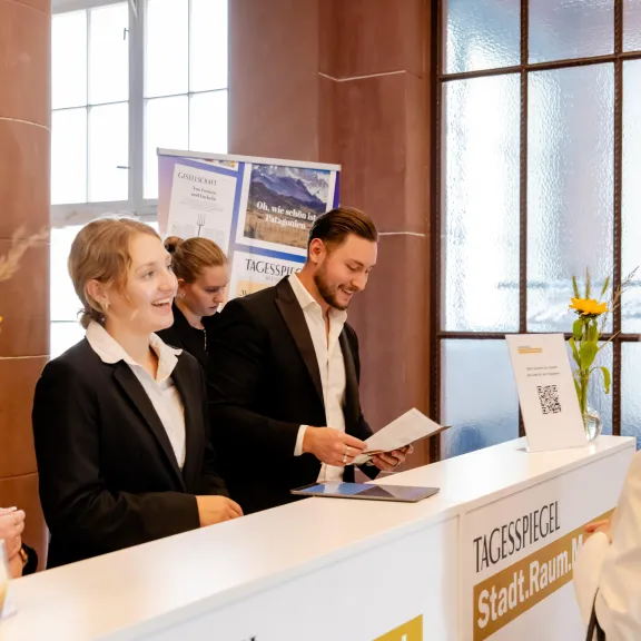 Mehrere Hosts und Hostessen in schwarzen Hosenanzügen stehen hinter der Akkreditierung und übergeben einer Dame einen Flyer – Eventpersonal und Promotionteam im Einsatz.