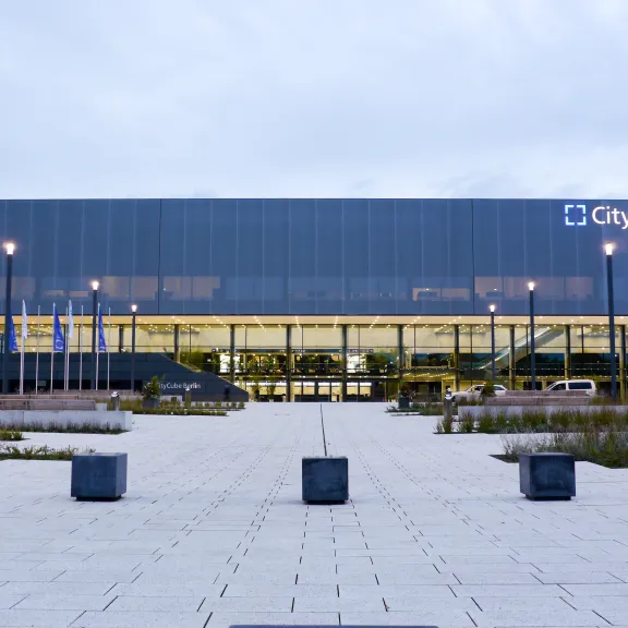 Moderne Glasfassade des CityCube Berlin bei Dämmerung, beleuchtet und umgeben von Parkplatz.