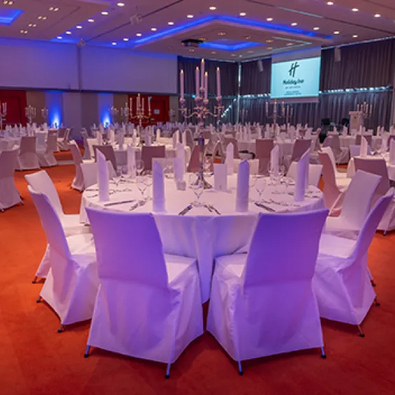 Eleganter Bankettsaal mit runden Tischen, weißen Stuhlhussen und blauer Beleuchtung.