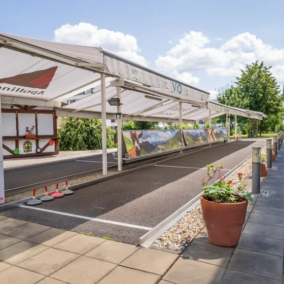 Überdachte Boule-Bahn mit seitlich angeordneten Pflanzkübeln und Sitzgelegenheiten.