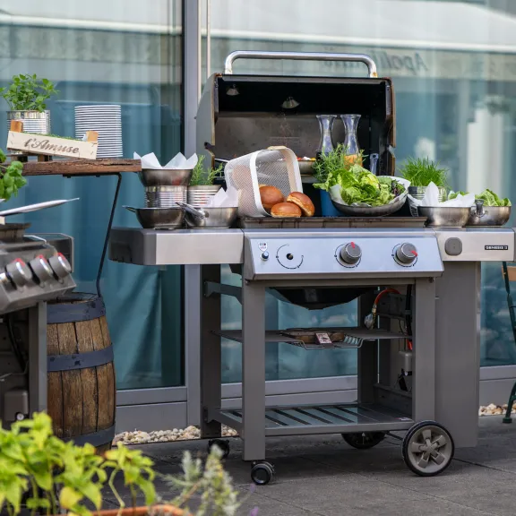 Grill mit frischem Gemüse, Brot und Kräutern auf Terrasse neben einem Tisch und Stühlen.