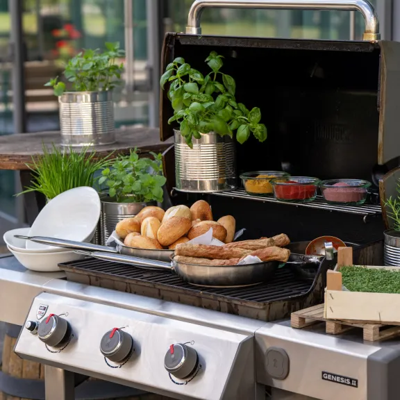 Eisstockpaket: Grill mit Brötchen, Würstchen, Kräutern und Saucen im Freien neben der Eisstockbahn.