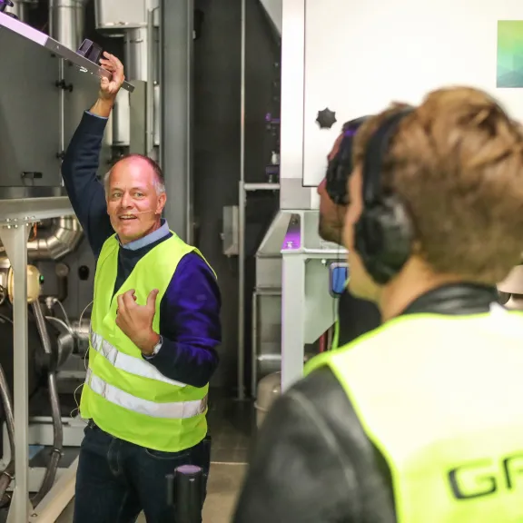 Mann in Warnweste erklärt Technik einem Besucher in Industrieumgebung.