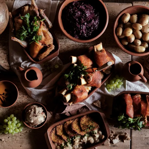 Rustikales Essen mit Braten, Kartoffeln, Brot, Trauben und Rotkohl auf Holztisch angerichtet.