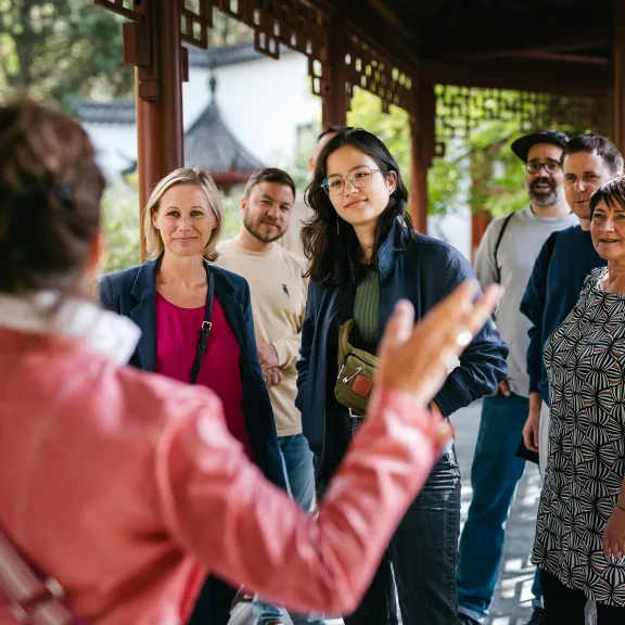 Gruppe hört einer Führerin in einem Garten aufmerksam zu, umgeben von Grün und Sonne.