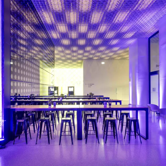 Moderner Raum mit langen Tischen, Hockern und lila Beleuchtung.