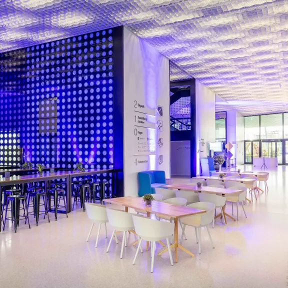 Modernes Café mit bunten Lichtern, hohen Tischen und weißen Stühlen, helle Atmosphäre.