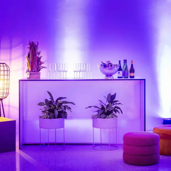 Bar mit Pflanzen, Getränken und Hockern, lila-blaues Licht, moderne Atmosphäre.