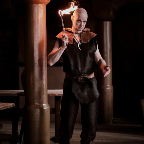 Mann in mittelalterlicher Kleidung hält Fackel in dunklem Raum