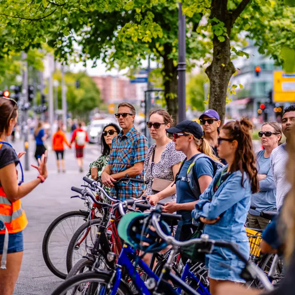 Gruppe von Radfahrer*innen hört einer Guide auf einer sommerlichen Stadtstraße zu.
