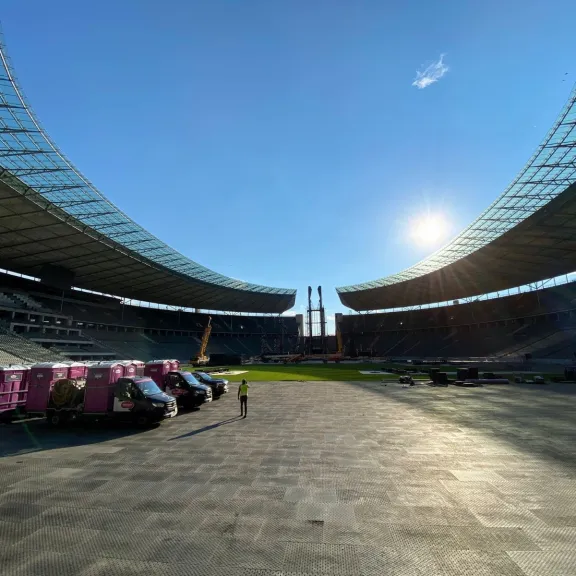 Auf diesem Bild sind mehere Servicefahrzeuge mit Anhänger zusehen, auf denen mehrere, pinke Wölkchen WC-Kabinen stehen. Diese Fahrzeuge stehen im Innenbereich des Berliner Olympiastadions. Der Himmel strahlt blau.