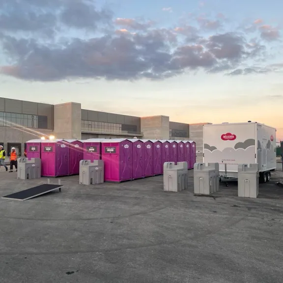 Auf einer großen Veranstaltungsfläche, auf der man im Hintergrund ein großes Firmengebäude sieht, stehen im Vordergrund viele, pinke Wölkchen WC-Kabinen. Daneben steht ein weißer WC-Wagen mit Wölkchen Logo und mehreren mobilen Handwaschstationen.