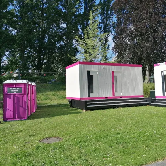 Auf einer Wiese stehen, im rechten Winkel zueinander, 2 weiße Wc-Conatiner mit pinkem Rahmen. Seitlich davin sieht man 2 pinke WC-Kabinen stehen.