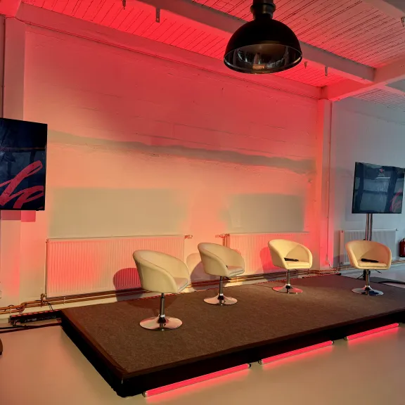 Kleine Bühne mit drei weißen Stühlen, roten Lichtern und zwei Bildschirmen.