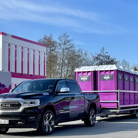 Hier wird ein Pick Up geuegt, der einen Anhänger zieht, auf dem mehrere, pinke Wölkchen WC-Kabinen stehen. Im Hintergrund ist ein WC-Wagen zu sehen, neben dem zwei, aufeinander stehende WC-Container, mit pinker Umrandung stehen.